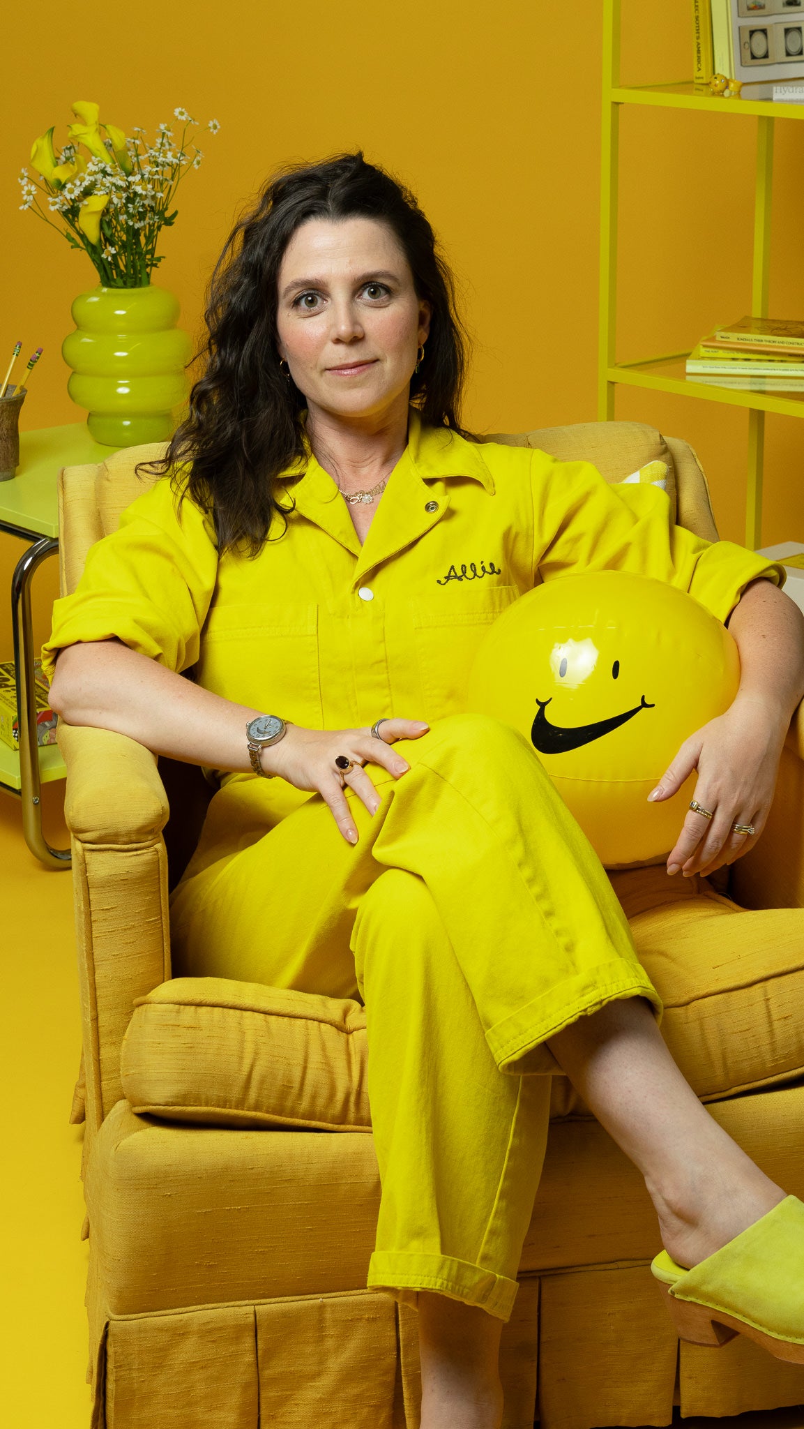 Allie Pisarro-Grant wearing a yellow jumpsuit with a smiley face ball, sitting on a yellow chair against a yellow background.