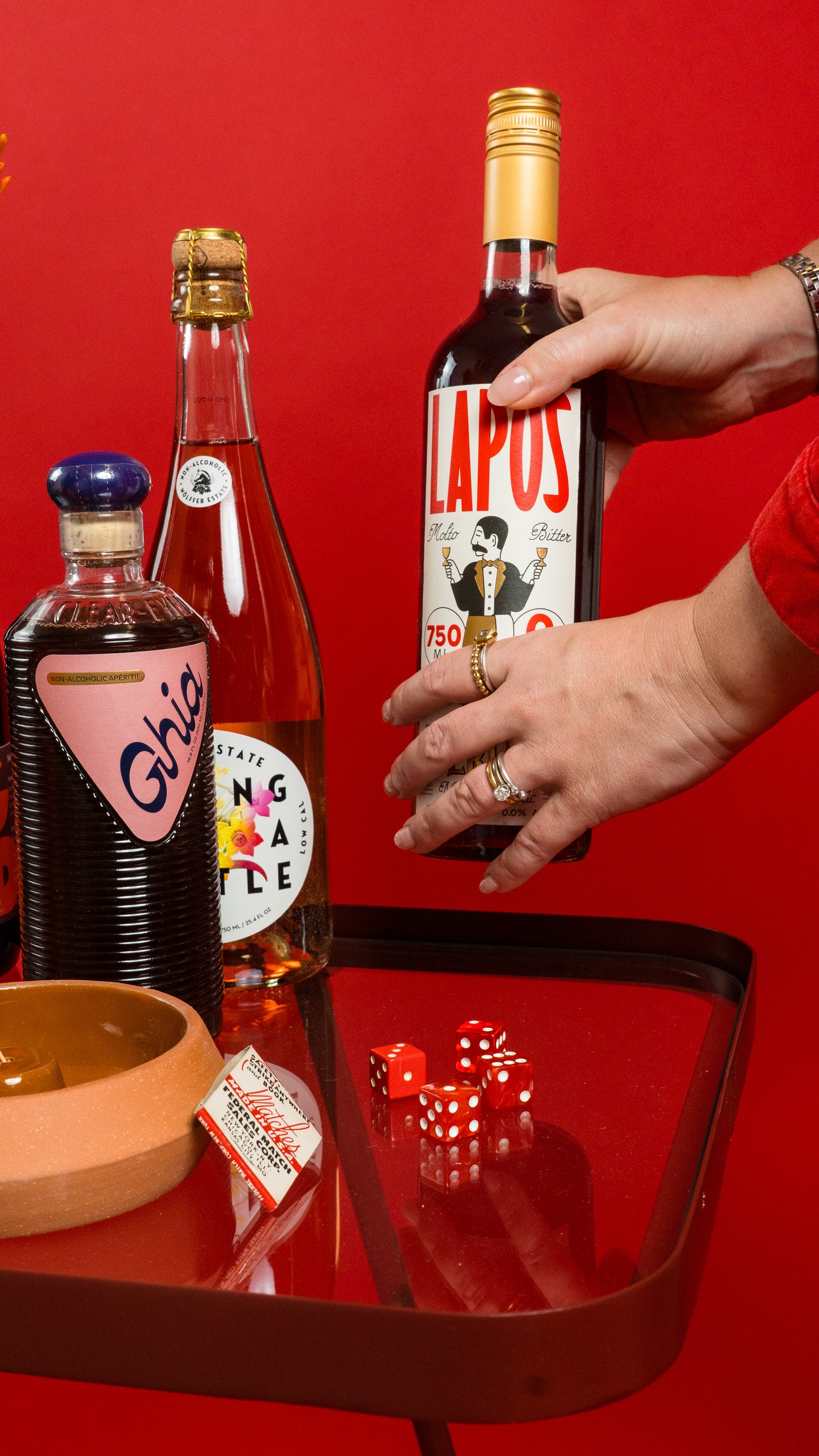 Person holding a bottle of Lapos against a red background with other bottles and dice on a table.