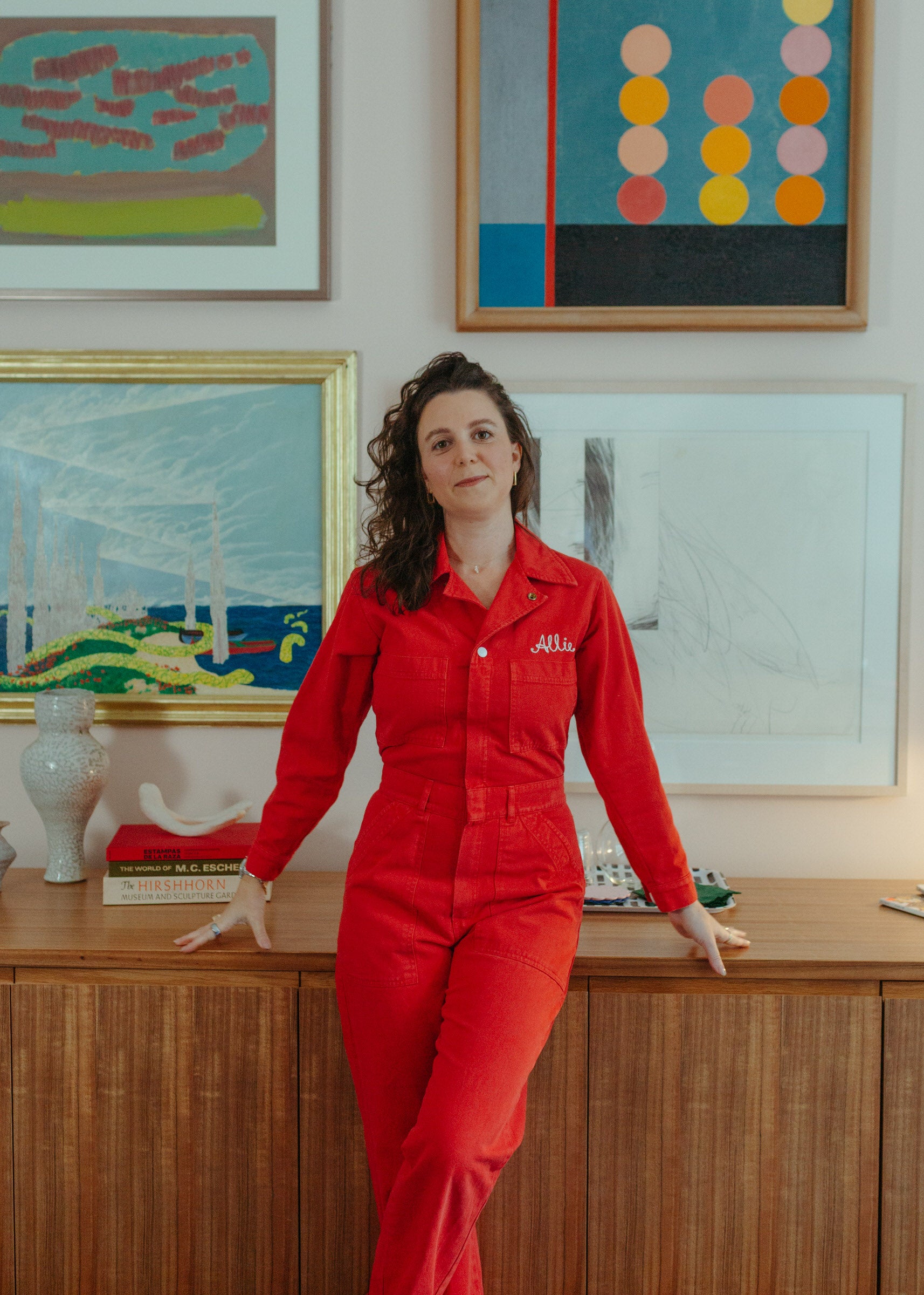 Allie Pisarro-Grant in a red jumpsuit standing in front of colorful abstract paintings on a wall.