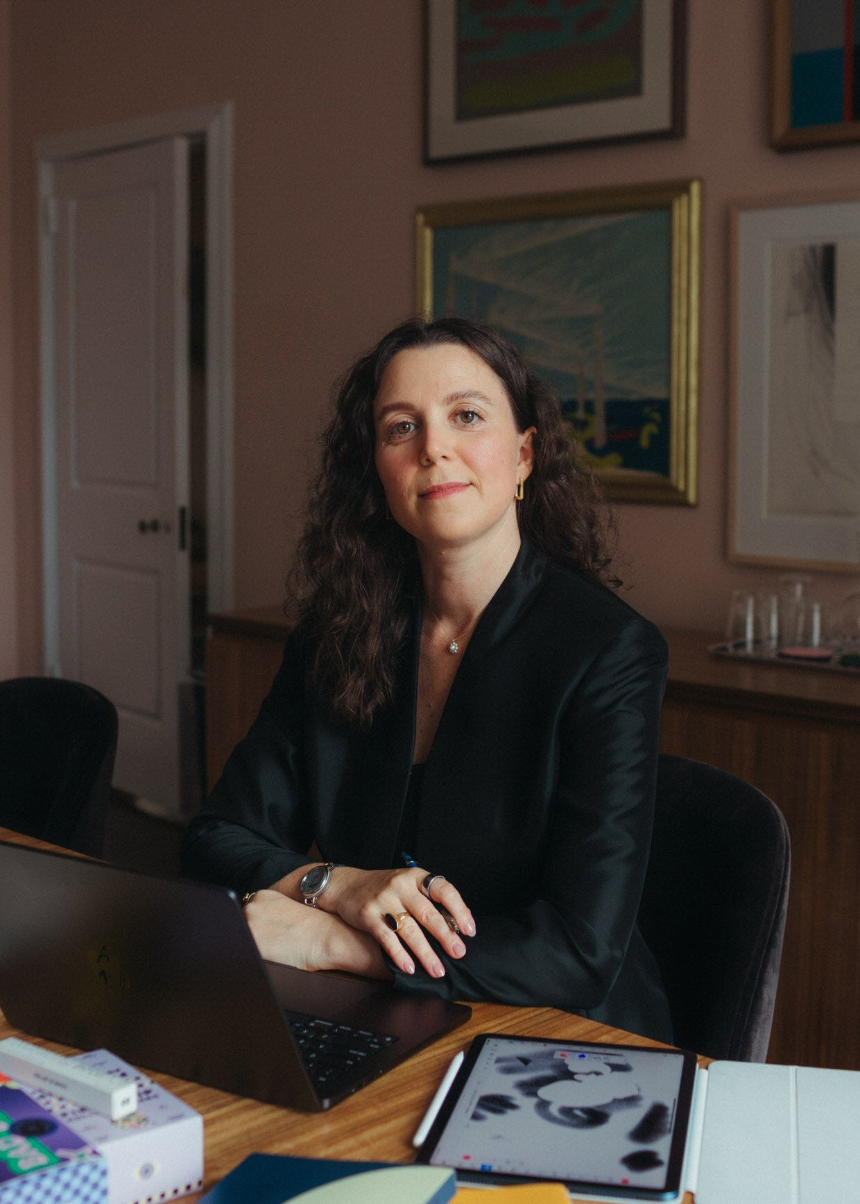 Allie Pisarro-Grant in the Hanes House board room at a wooden desk, wearing a black blazer. She is slightly smiling with a laptop in front of her, surrounded by notebooks, a tablet, and framed artwork on a dusty pink wall.
